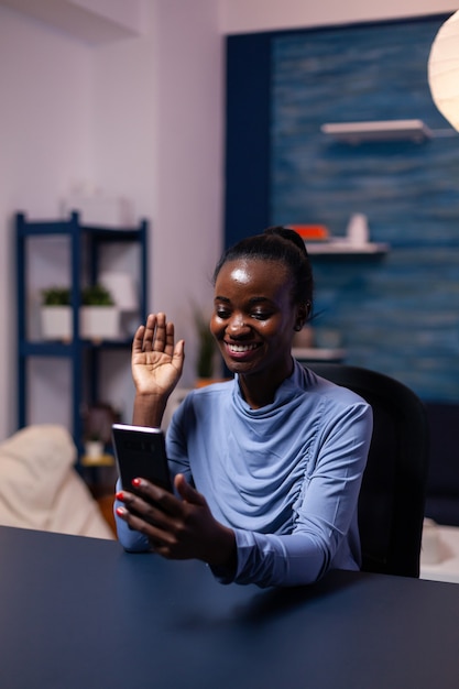 Vrouw met een donkere huid zegt hallo tijdens een videoconferentie die 's avonds laat op kantoor aan het bureau zit. zwarte freelancer die werkt met een team op afstand dat virtuele online conferentie chat.