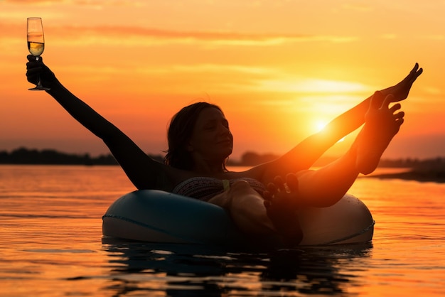 Foto vrouw met een glas champagne liggend op opblaasbare ring in water bij zonsondergang of zonsopgang