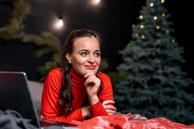 Vrouw met een glimlach op de rode deken Een dame met een dromerige glimlach liggend op de plaid met een laptop aan haar zijde Liggend meisje met een notitieboekje bij de kerstboom