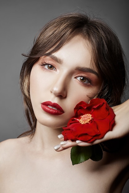 Vrouw met een grote rode bloem in haar haar. Bruinharig meisje met een rode bloem die zich voordeed op een grijze muur. Grote mooie ogen en natuurlijke make-up