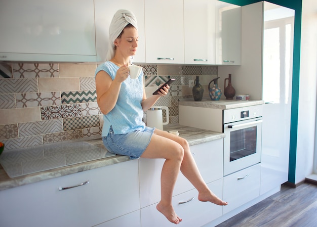 Vrouw met een handdoek op haar hoofd in haar keuken die ontbijt heeft en naar de telefoon kijkt