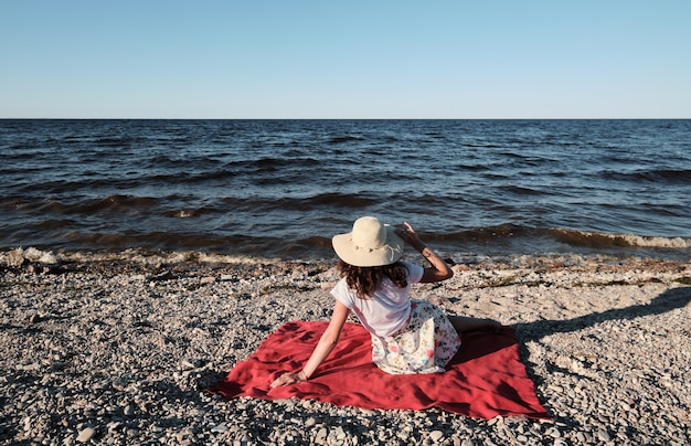 Vrouw met een hoed zit op een rode deken op het strand en kijkt naar het water