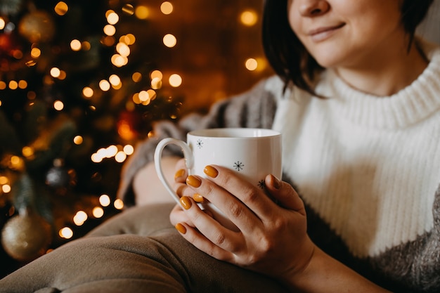 Vrouw met een kopje koffie of thee, zittend op de bank naast een kerstboom thuis