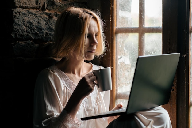Vrouw met een laptop en een kopje koffie bij het raam Vrouw van middelbare leeftijd in een wit overhemd Ochtendkoffie Freelancer