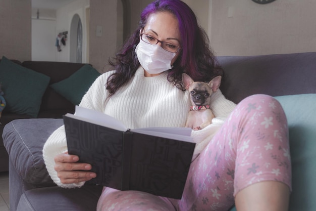 Vrouw met een masker die een boek leest naast haar huisdier een chihuahuahond die ze thuis in quarantaine zit vanwege de wereldwijde pandemie