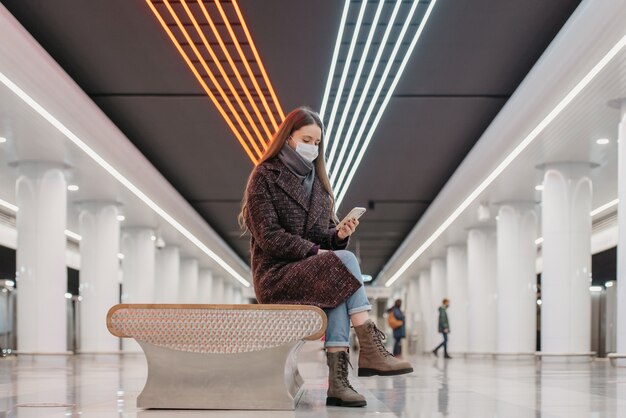 Vrouw met een medisch gezichtsmasker zit in het midden van het grote metrostation met een smartphone en leest het nieuws. Een meisje met lang haar met een chirurgisch masker houdt sociale afstand in de metro