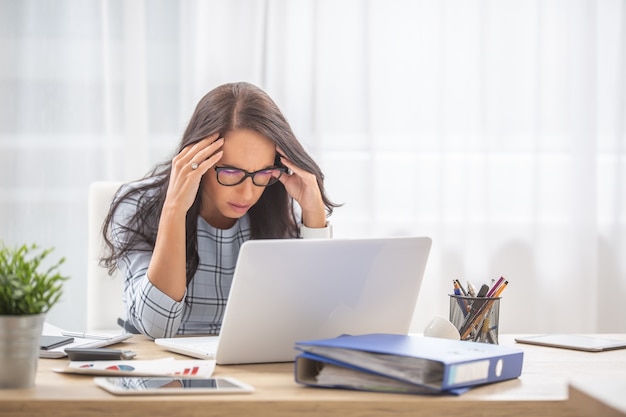 Vrouw met een migraine die het hoofd in haar handen houdt met gesloten ogen aan het bureau, voor de computer.