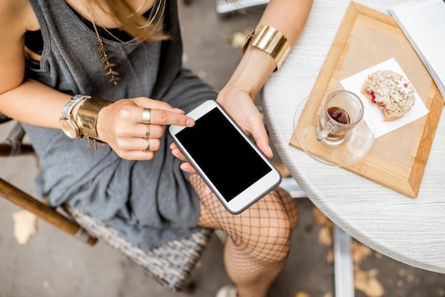 Vrouw met een smartphone met een leeg scherm die buiten in het café zit met cake en koffie op tafel