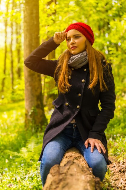 Vrouw met een zwarte jas en een rode hoed die geniet in een herfstpark, zittend op een boom glimlachend bij zonsondergang, verticale foto