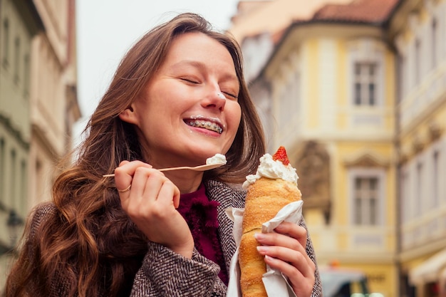 Vrouw met eetlust eet een traditionele Tsjechische zoete Trdelnik met vanilleroom en aardbeien met behulp van eco houten lepel in Praag straat