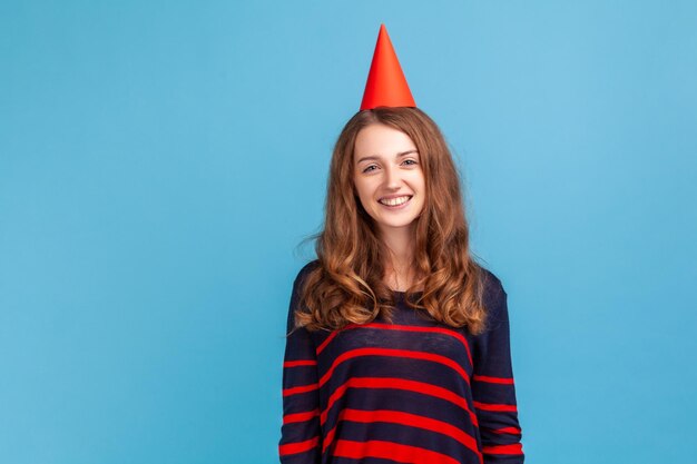 Vrouw met feestelijke stemming dragen gestreepte casual stijl trui staande gelukkige gezichtsuitdrukking viert haar verjaardag met partij kegel op haar hoofd Indoor studio shot geïsoleerd op blauwe achtergrond