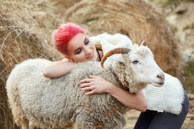 Vrouw met felrode make-up op haar gezicht knuffelt een gehoornde ram. creatieve hete roze make-up op meisjesgezicht, haarkleuring. portret van een meisje met een schaap. loop in het herfstbos. herfst wollen kleding