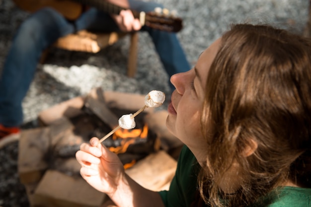 Vrouw met gebakken marshmallows en man met gitaar dicht bij kampvuur