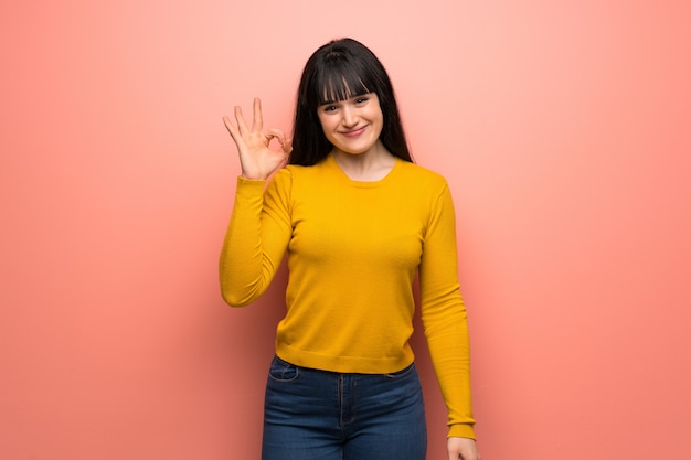 Vrouw met gele sweater over roze muur die ok teken met vingers toont