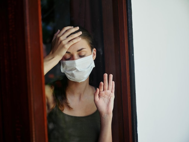 Vrouw met gesloten ogen in medisch masker die uit de raamvergrendeling kijkt