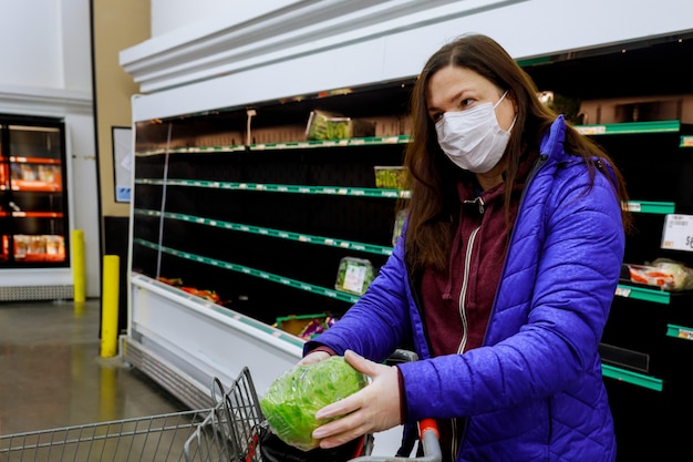 Vrouw met gezichtsmasker het kopen sla bij supermarkt met lege planken.