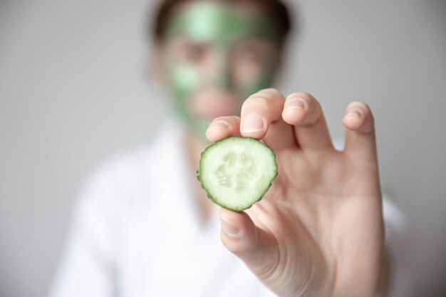 Vrouw met groen masker op haar gezicht die verse komkommerplak vage achtergrond houdt