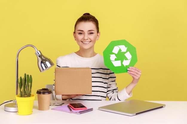 Vrouw met groen recyclingbord in de hand en kartonnen pakket zittend op de werkplek met laptop