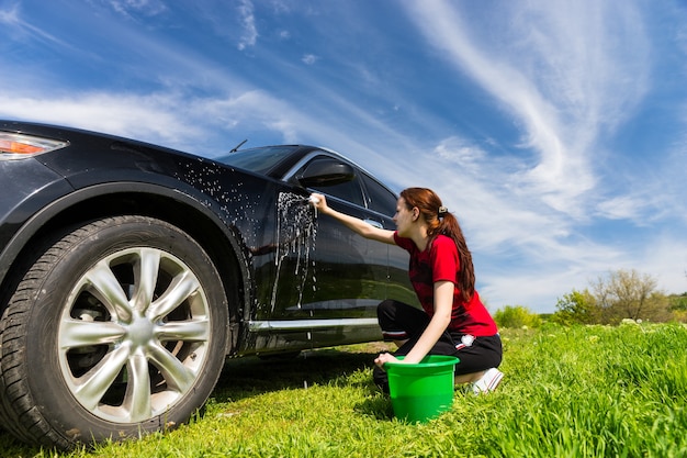 Vrouw met groene emmer wassen zwarte luxe voertuig met zeepachtige spons in groen veld op zonnige dag met blauwe lucht