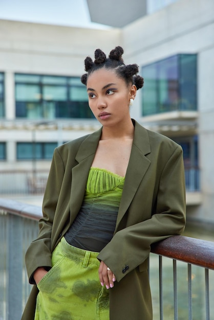 Vrouw met haarbroodjes gekleed in modieuze groene kleding poses op balkon houdt hand in zak diep in gedachten. trendy hipster meisje