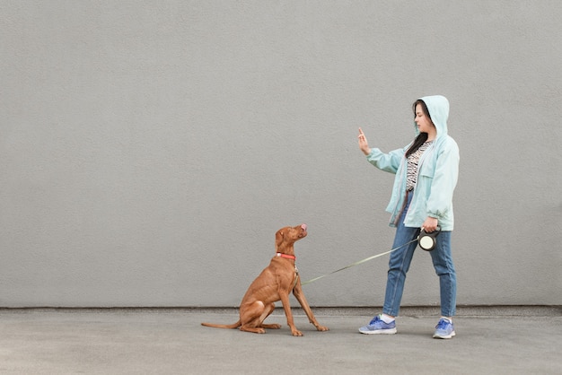 Vrouw met jas traint een hond tegen een grijze muur