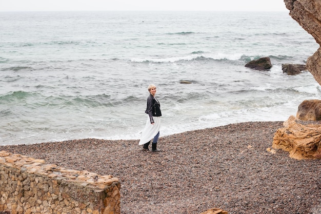 Vrouw met jurk en jas die in de herfst in de buurt van zee loopt