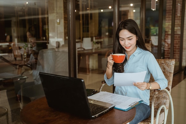 Vrouw met koffiekopje papierwerk lezen tijdens het gebruik van laptop in co-working space