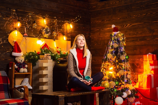 Vrouw met kop og warme drank in haar handen wachtend op Nieuwjaar. Gelukkige jonge mooie dame bij de open haard in de buurt van de kerstboom.