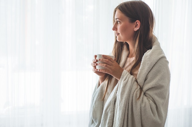 Vrouw met kop warme drank bij het raam. kijkend naar het raam en drink thee. goedemorgen met thee. herfst wintertijd