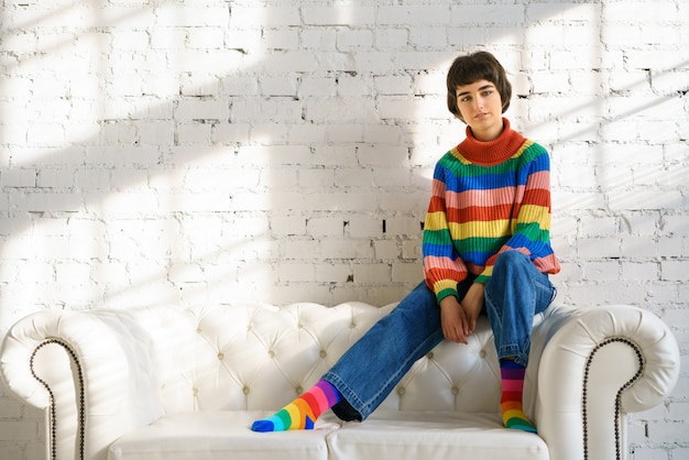 vrouw met kort haar in een regenboog trui en sokken zit op een witte sofa, het concept van seksuele minderheden