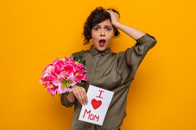 Vrouw met kort haar met wenskaart en boeket bloemen kijkend naar camera verbaasd en verrast vieren internationale Vrouwendag 8 maart staande over oranje achtergrond