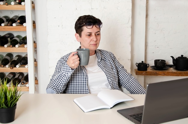 Vrouw met kort zwart haar met een bril drinkt koffie terwijl ze aan het bureau op kantoor zit, voor een opengeklapte laptop. vrouw drinkt thee, plant zaken, maakt aantekeningen in notitieblok. Thuiskantoor concept
