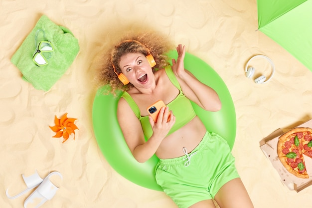 vrouw met krullend borstelig haar roept uit vreugde krijgt uitstekend nieuws houdt moderne smartphone gekleed in zomerkleren poses op strand heeft goede rust