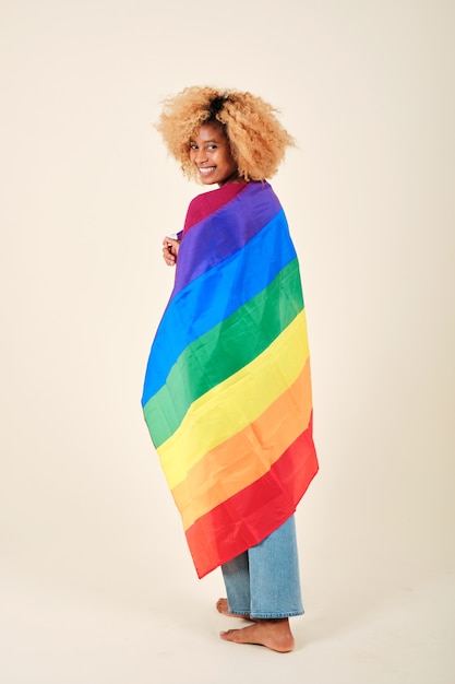 Vrouw met krullend haar die in de camera kijkt en glimlacht terwijl ze is omwikkeld met een lgbt-trotsvlag op een geïsoleerde achtergrond. LGBTQ-gemeenschapsconcept.