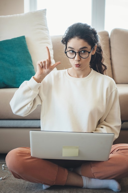 Vrouw met krullend haar gebaart het idee-teken terwijl ze online lessen op de vloer op laptop heeft