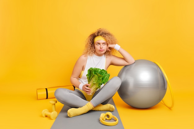 vrouw met krullend haar leunt naar zwitserse bal houdt groene groente zit gekruiste benen op fitnessmat voelt zich moe na de training omringd door fitball hoelahoep halters karemat
