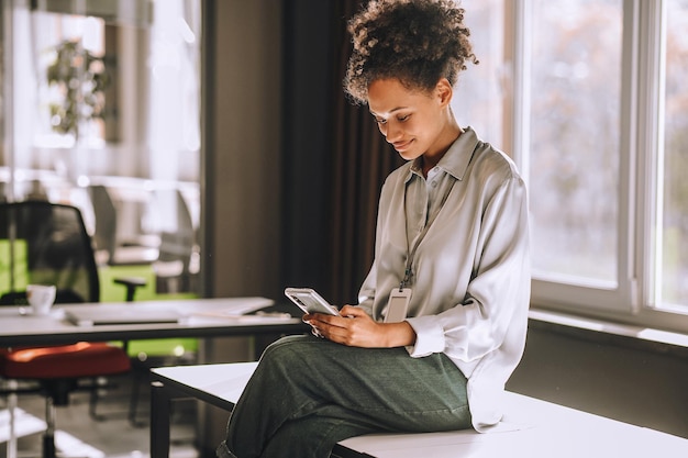 Vrouw met krullend haar zittend op de tafel met een telefoon in handen