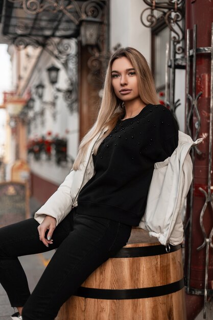 vrouw met lang blond haar in een trendy wit jasje zit op een houten vintage vat