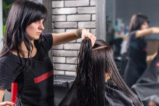 Vrouw met lang nat haar die op kapsel wacht terwijl vrouwelijke kapper haar haar kamt, zittend in een fauteuil in schoonheidssalon