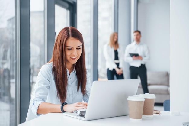 Vrouw met laptop die voor een groep jonge succesvolle teams zit die binnenshuis op kantoor werken en communiceren