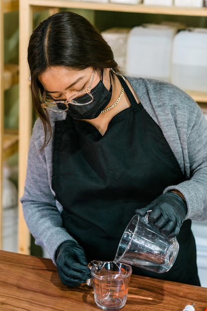 Vrouw met masker en handschoenen die vloeistof meten in een glazen beker om essentie te maken.