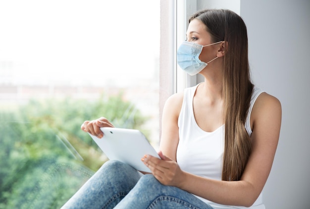 Vrouw met medisch masker geïsoleerd Blijf thuis Meisje met gezichtsmasker Gebruik tablet Werkstudie bij vensterbank