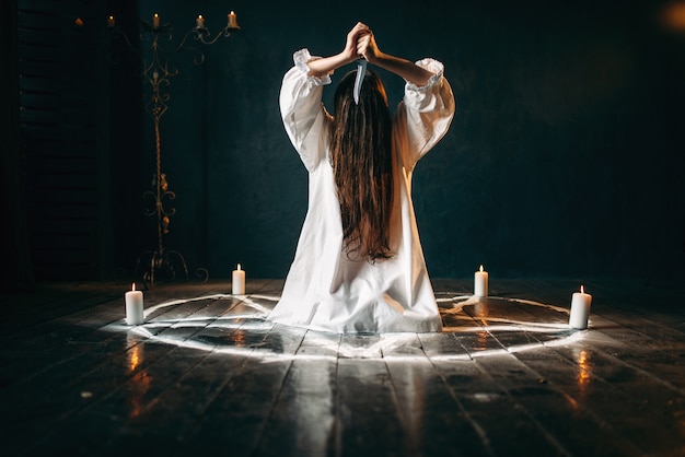 Vrouw met mes in handen die in pentagramcirkel met kaarsen zitten. Donker magisch ritueel, occultisme en exorcisme, bovennatuurlijke kracht