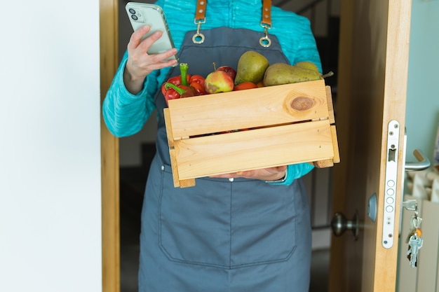 Foto vrouw met mobiele telefoon in de hand en houten kist met verse groenten en fruit.