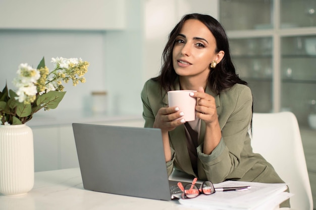 Vrouw met mok met behulp van laptop in de keuken van het appartement