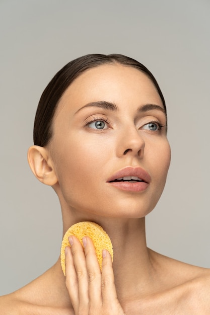 Vrouw met naakte make-up en blote schouders die haar nek schoonmaakt met een exfoliërende spons geïsoleerd