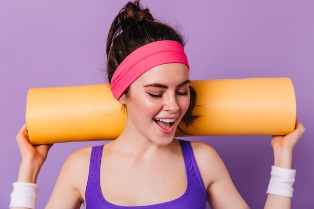 Vrouw met oranje fitnessmat op paarse achtergrond Brunette in hoofdband glimlacht