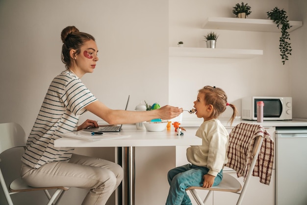 Vrouw met plekken onder de ogen die voor de laptop zit en een lepel pap geeft aan het meisje aan de keukentafel