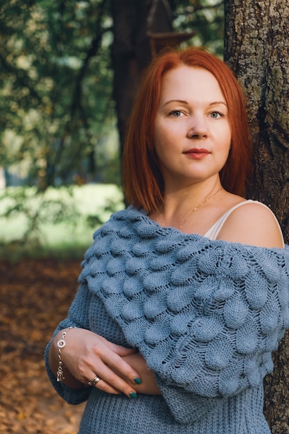 Vrouw met rood haar en een gebreide trui staat bij een boom in het park in de stralen van de ondergaande zon