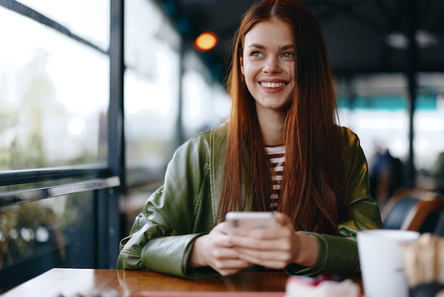 Vrouw met rood haar freelance blogger zittend in café met telefoon en glimlachend hipster influencer meisje in modieuze lifestyle kleding